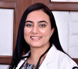 The image shows a woman wearing a white lab coat, standing against a wall with a brick pattern, smiling at the camera.