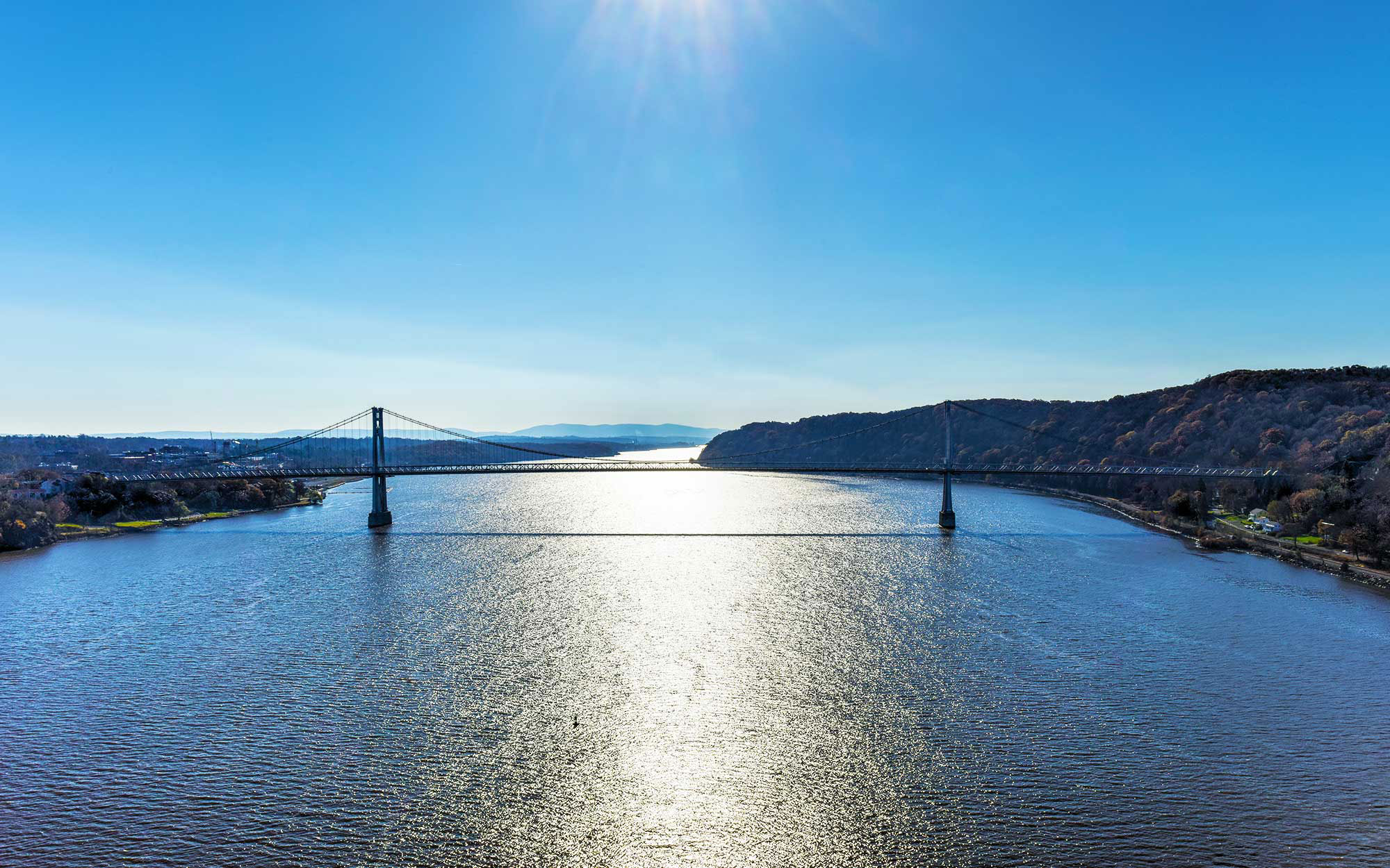 The image shows a tranquil scene with a large body of water, possibly a river or lake, under a clear blue sky. A bridge spans across the water, connecting two land masses. The perspective of the photo suggests it was taken from an elevated position, providing a panoramic view of the landscape. The image conveys a sense of calm and natural beauty.