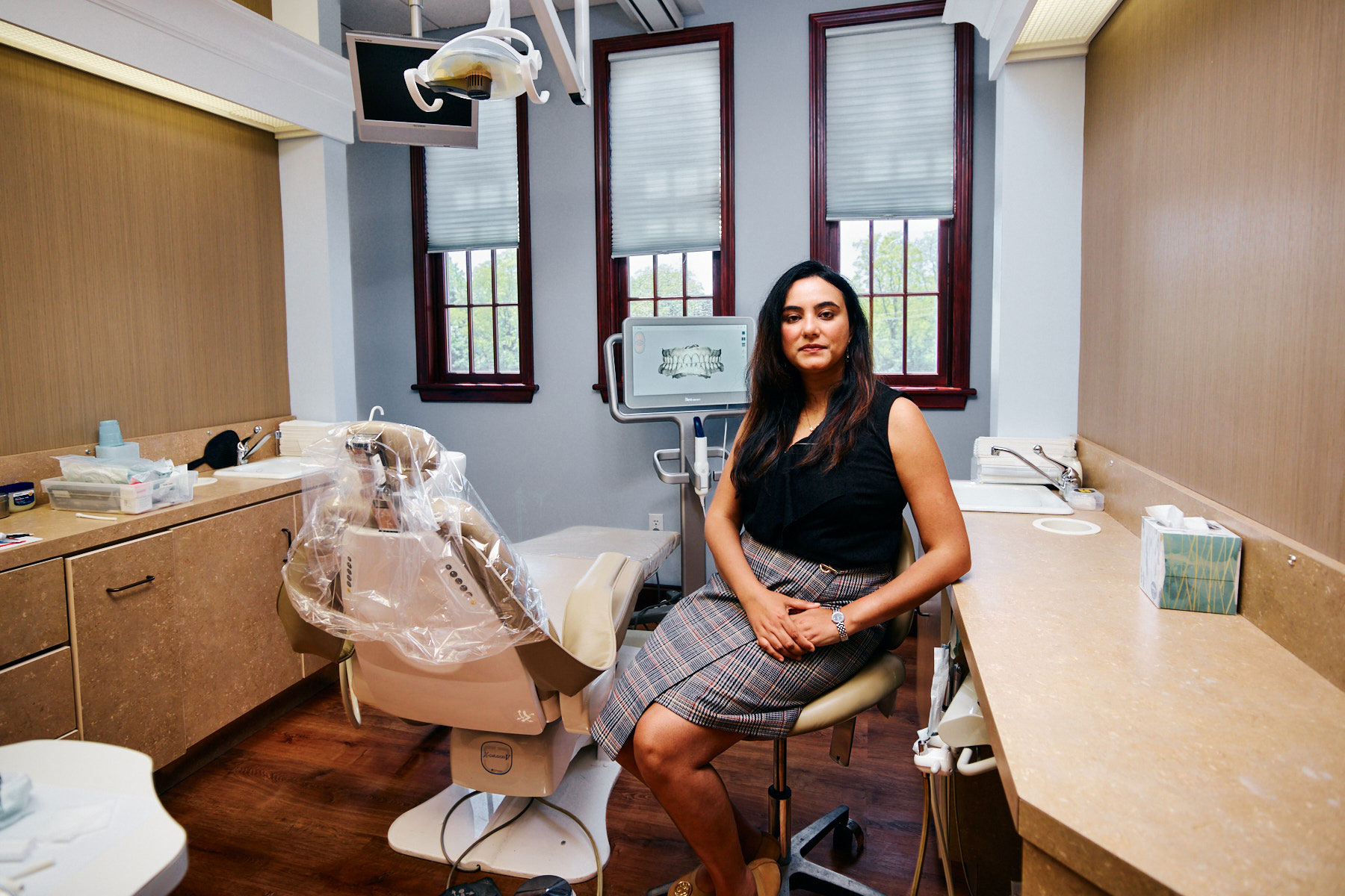 A woman sitting at a dental office chair, smiling towards the camera, with dental equipment in the background.