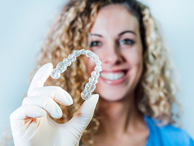 A woman wearing a face mask holds up a transparent retainer with a smile on her face.