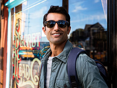 The image depicts a smiling man wearing sunglasses, standing outside a shop with a sign that reads Secret Garden. He has a backpack and is dressed in casual attire.