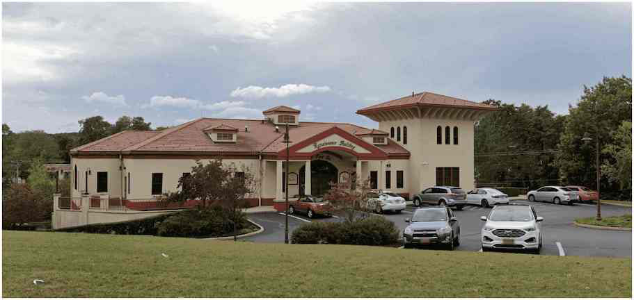 The image shows a two-story building with a prominent front entrance, featuring a covered porch and a red roof. There are cars parked in front of the building, which appears to be a residential structure.